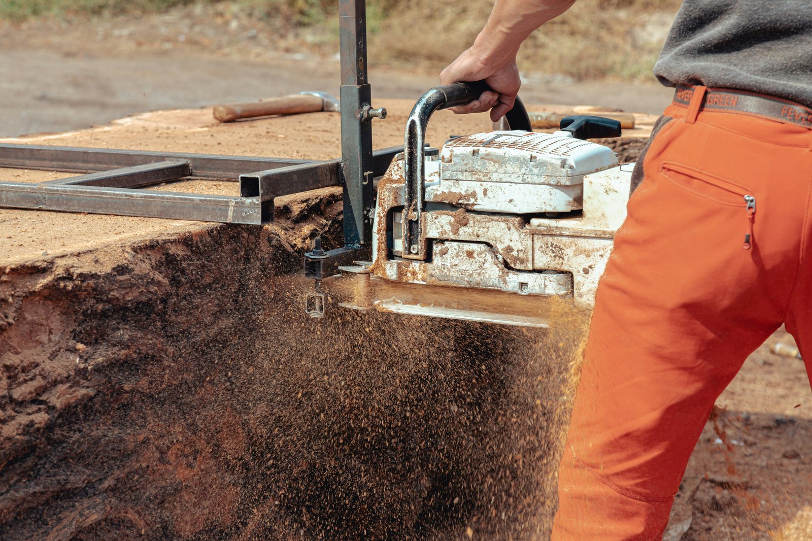 A person is using a chainsaw to cut through a large piece of wood, creating a cloud of sawdust. The individual is wearing orange pants and a grey shirt, and they are outdoors in a rustic setting. The chainsaw is mounted on a metal frame to guide the cut, and the scene captures the dynamic action of cutting wood.